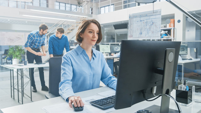 In the Industrial Engineering Facility: Portrait of the Smart and Beautiful Female Engineer Working on Desktop Computer. In the Background Specialists and Technicians Working with Drafts and Drawings