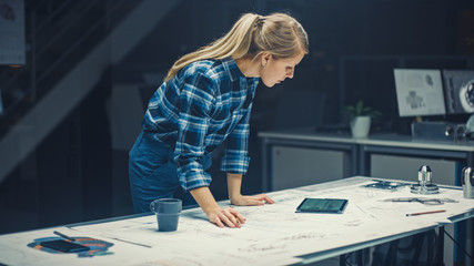 In the Dark Industrial Design Engineering Facility: Female Engineer Works with Blueprints Laying on a Table, Uses Digital Tablet and Drinks Coffee. On the Desktop Drawings and Engine Components