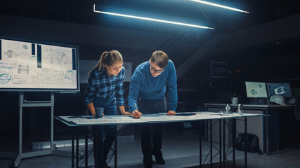 In the Dark Industrial Design Engineering Facility Male and Female Engineers Talk and Work on a Blueprints Using Digital Tablet and Conference Table. On the Desktop Drawings and Engine Components