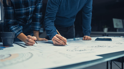 Close-up Shot with Focus on Hands of Male and Female Engineers Work on a Blueprints Using Pencils on Conference Table. In the Dark Industrial Design Engineering Facility.