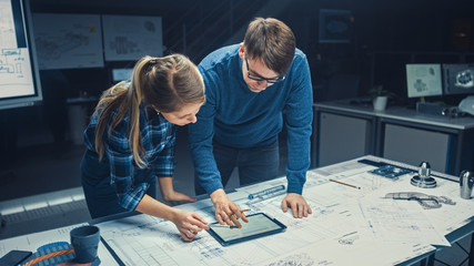 In the Dark Industrial Design Engineering Facility Male and Female Engineers Talk and Work on a Blueprints Using Digital Tablet and Conference Table. On the Desktop Drawings and Engine Components
