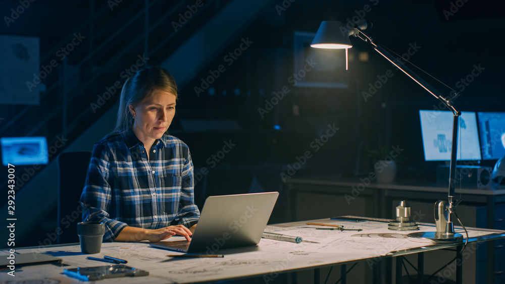Poster beautiful female engineer sitting at her desk works on a laptop computer. blueprints lying on a tabl