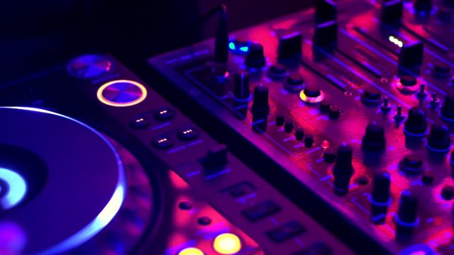 Turntables, buttons, sliders and wires on sound mixing controller deck under multicolor party lighting, close-up. Disc jockey mix tracks, play electronic music on pro audio equipment at night club.
