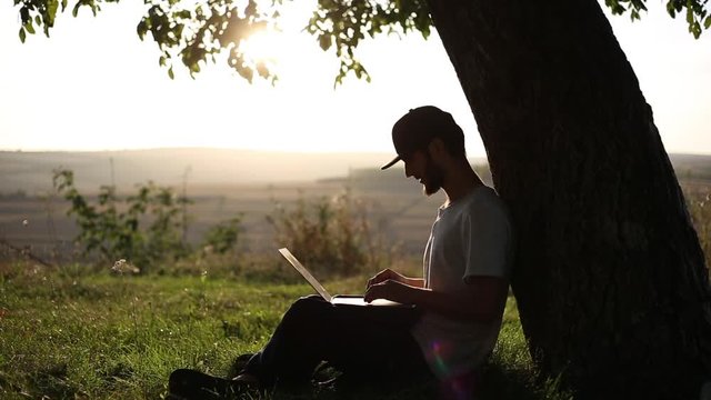 Young hipster sitting on grass in park, lean against big tree with laptop on legs, typing, working. Beautiful background of sunset and small mountains. Wearing cap.