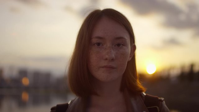 Portrait shot with tracking of face of freckled teenage girl with red hair posing for camera at sunset