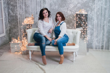 Obraz premium Two beautiful girls celebrates Christmas, New Year. Girls in the New Year's interior of the room are sitting on a white sofa against the background of garlands