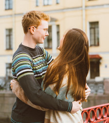 Girl with long thick dark hear embracing redhead boy in the blue t-shirt on a bridge, young couple. Concept of teenage love and first kiss, sincere feelings of man and woman