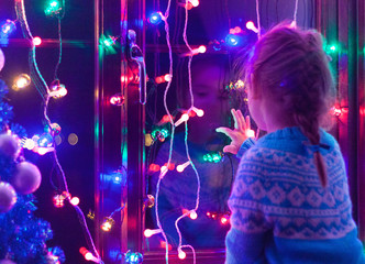Happy little cute, beautiful girl 5 years old is sitting on the window and playing with Christmas lights and  Christmas decoration. Dark-blue theme. 