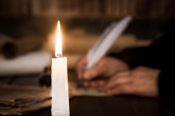A lit candle against the background of blurred scrolls, writer's hand, books and bird's feather