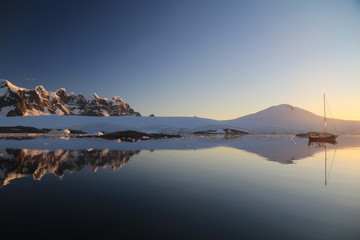 ośnieżone skalne wybrzeże antarktydy odbijające się w gładkiej tafli wody o zachodzie słońca