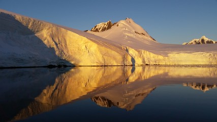 ośnieżone skalne wybrzeże antarktydy odbijające się w gładkiej tafli wody o zachodzie słońca