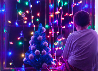 Teenager boy is sitting on the window and looking at the Christmas lights