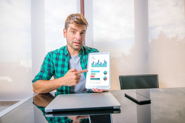Young business man sitting at office desk looking at camera and pointing at a tablet with financial information displayed in graphical form (column graph, stacked bar graph and pie graph). Screen gra