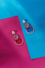 Subject shot of stud earrings in the form of a shimmering pearl with dangling various rings and large rainbow paillettes. The earrings are isolated on the blue and pink surface with geometric design.