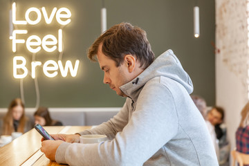 Alone sad unhappy man sitting in cafe with smartphone and cup of cappuccino with neon sign love feel brew on background. Man with broken heart. Concept of modern loneliness people.