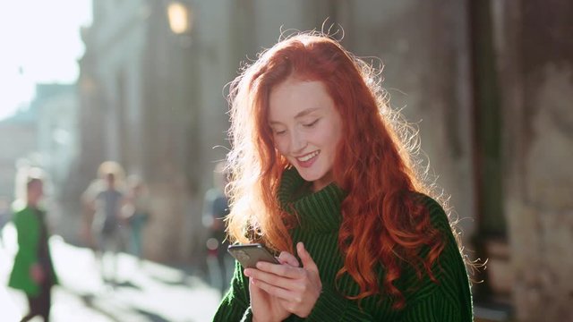 Smiling young woman with red hair using her phone chatting with friends in the city center feel happy positive emotion