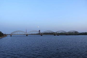 Long metal railroad arched bridge across the Daugava river in Riga against stand alone building, TV tower and bright moon forward view on sunsetsky