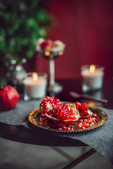 Still life composition with juicy red cut cleared pomegranate on a copper plate, burning candles and other oriental decor on the black table. Soft selective focus. Vertical card. Copy space.