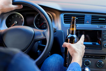 Closeup male hand is holding beer behind steering wheel of car. Drunk driver is riding in...