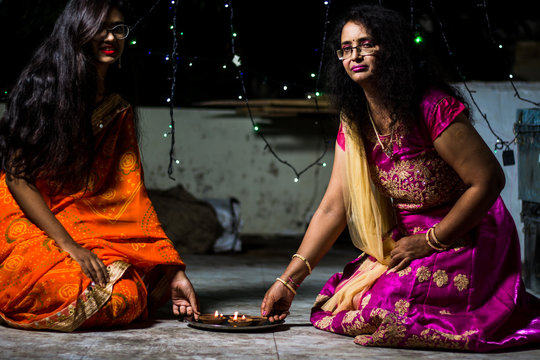 Image of An young and beautiful Indian Bengali woman in Indian traditional  dress is lighting Diwali diya/lamps sitting on the dark floor indoor to  celebrate Diwali. Indian lifestyle and Diwali celebration-AK715317-Picxy