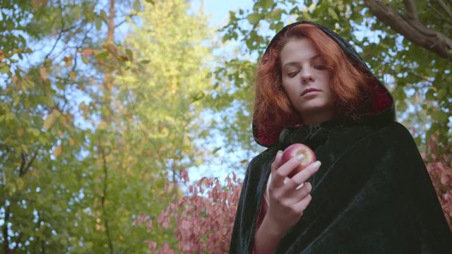 Close-up of attractive red-haired caucasian woman in a black gown looking at the apple in her hands. Fairy girl standing in the autumn forest. Snow White style.