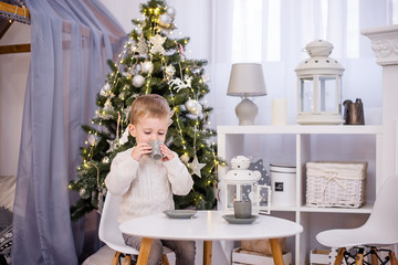 A little boy in a white pullover plays tea, drinks cocoa from a gray cup at his white table in the children's room. In the background there is a Christmas tree with garlands of lights. Copy space 