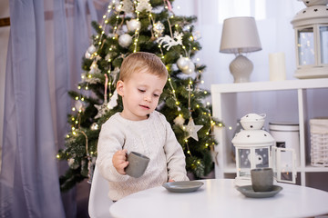 A little boy in a white pullover plays tea, drinks cocoa from a gray cup at his white table in the children's room. In the background there is a Christmas tree with garlands of lights. Copy space 