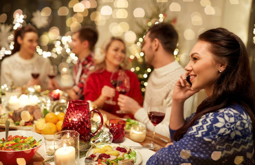 holidays, communication and celebration concept - happy young woman calling on smartphone and having christmas dinner with friends at home