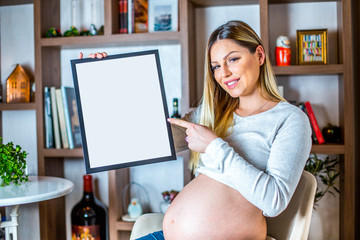 Young beautiful pregnant girl holding white empty board in her home during daytime