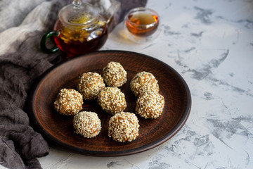On a brown clay plate are Indian sweets Lada. They are sprinkled with sesame seeds. In the background is flower tea with ginger and lime. Everything is located on a gray background decorated in granit