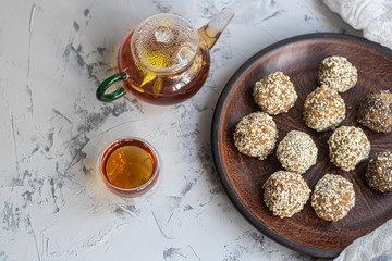 On a brown clay plate are Indian sweets Lada. They are sprinkled with sesame seeds. In the background is flower tea with ginger and lime. Everything is located on a gray background decorated in granit