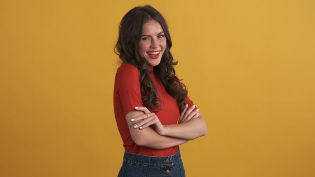 Pretty brunette girl in red top with hands crossed charmingly winking in camera over yellow background