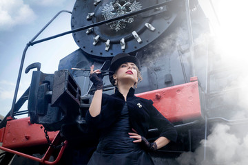Steampunk girl in a black dress and hat near an old steam locomotive and large iron wheels. Blond beauty. Vintage portrait of the last century, retro journey.