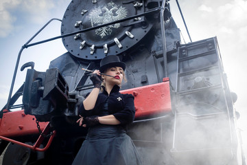 Steampunk girl in a black dress and hat near an old steam locomotive and large iron wheels. Blond beauty. Vintage portrait of the last century, retro journey.