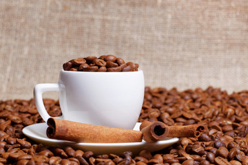 Coffee cup demitasse full of coffee beans and cinnamon sticks in the heap coffee beans on burlap background