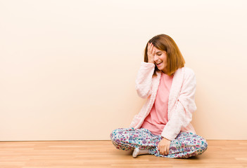 young woman wearing pajamas sitting at home laughing and slapping forehead like saying d‚Äôoh!...