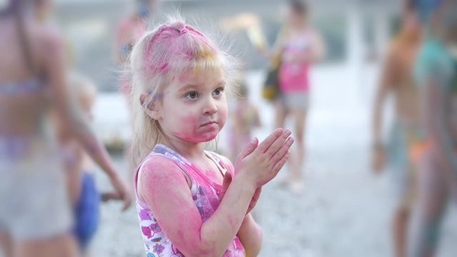 A cute little girl participates in a paint festival by playing with colorful paints and sand. His face and clothes were stained with bright colors.