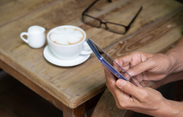 Hands hold and using smartphone during have a cup of coffee with latte art, eyeglasses on wooden table in coffee shop.