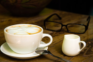Top view of white coffee cup with latte art, little milk mug and eyeglasses on wooden table in coffee shop.