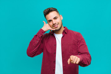 young hispanic man smiling cheerfully and pointing to camera while making a call you later gesture,...