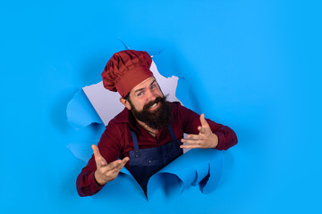 Bearded man in chef uniform. Professional happy man chef looking through paper hole. Cooking, profession and business. Professional approach to business. Male chef, cook or baker in hat and apron.