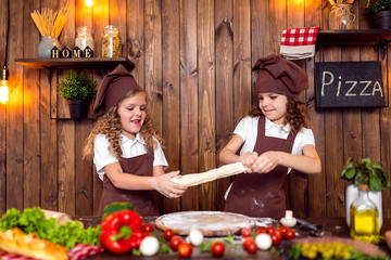 Happy little girls with rolling pin and spatula smiling and looking at camera while preparing delicious pizza in cozy kitchen together