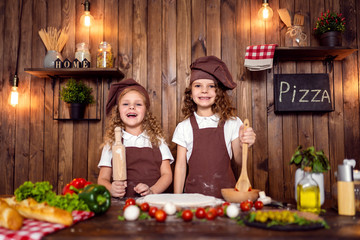Happy little girls with rolling pin and spatula smiling and looking at camera while preparing delicious pizza in cozy kitchen together