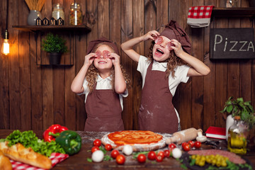 Delighted little sisters in aprons and hats holding pieces of sausage near eyes while preparing...
