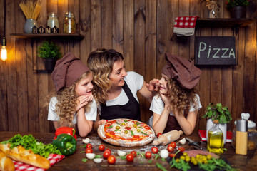 Cheerful woman with cute girls in aprons and hats smiling and looking each other during pizza preparation in kitchen at home