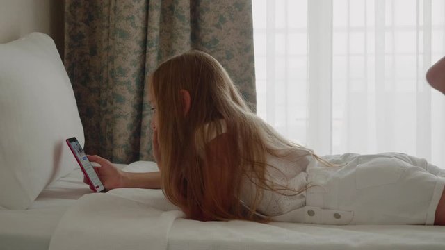 Teenage girl using smartphone in bedroom. Side view of adorable teen girl with beautiful long hair lying on bed and using mobile phone. Digital device concept