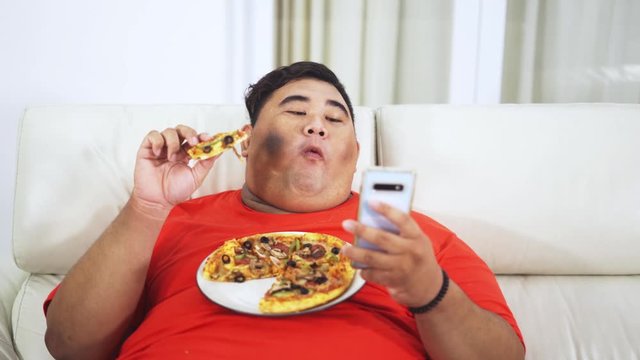 Overweight man eating a plate of pizza while using mobile phone on the sofa in the living room at home. Shot in 4k resolution