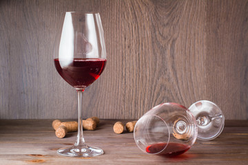 One glass with the remains of red wine is lying, the other is standing filled with red wine and cork on a wooden table