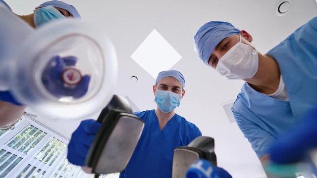 Three doctors holding medical instruments in the emergency room. Medical specialists in blue uniform with masks using medical tools to give resuscitation to a patient in clinic.