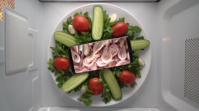 Cell phone on plate with boiled shrimp on screen in microwave. Food blog concept. Top view of mobile phone with food footage on screen rotating in microwave oven.